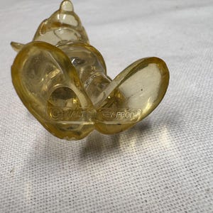 K&ouml;nnte beinhalten: Durchscheinende gelbe Figur einer Zeichentrickfigur, die auf dem R&uuml;cken liegt. Die F&uuml;&szlig;e der Figur sind nach oben gerichtet. Die Worte "&copy; WALT DISNEY PROD" sind auf dem Fu&szlig; zu sehen. Hergestellt aus transparentem Material.