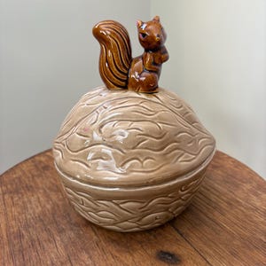 May include: A ceramic nut-shaped container with a brown squirrel figurine on top. The container is tan with a textured surface, and the squirrel is brown with a fluffy tail. The container is sitting on a wooden surface.