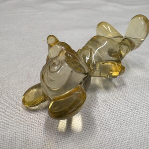 K&ouml;nnte beinhalten: Eine klare, bernsteinfarbene Glasfigur eines liegenden Tieres, m&ouml;glicherweise ein Kaninchen oder eine &auml;hnliche Kreatur. Die Figur liegt auf dem R&uuml;cken, die Pfoten und Ohren ausgestreckt. Das Glas hat eine glatte, gl&auml;nzende Oberfl&auml;che.