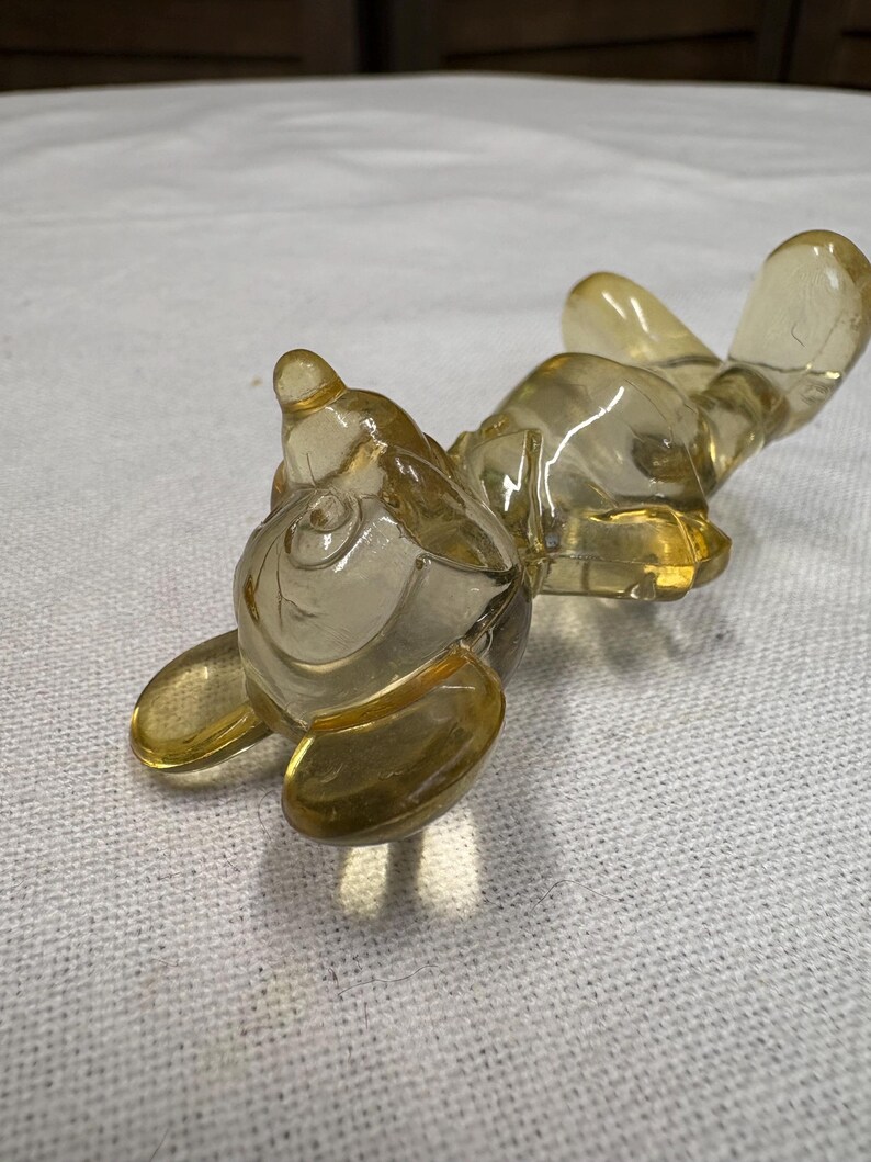 K&ouml;nnte beinhalten: Eine klare, bernsteinfarbene Glasfigur eines liegenden Tieres, m&ouml;glicherweise ein Kaninchen oder eine &auml;hnliche Kreatur. Die Figur liegt auf dem R&uuml;cken, die Pfoten und Ohren ausgestreckt. Das Glas hat eine glatte, gl&auml;nzende Oberfl&auml;che.