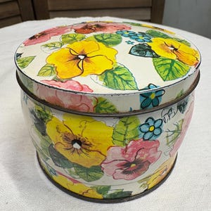 May include: A round, vintage tin box with a lid. The box is painted with a floral design featuring yellow, pink, and blue flowers with green leaves against a white background. The tin has a dark metal rim.