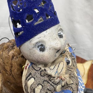 May include: A handmade doll with a painted face, wearing a blue velvet hat with cut-out designs. The doll's clothing features a patterned fabric with blue, black, and cream colors. The doll is positioned on a wooden surface.