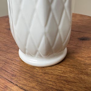 May include: A white ceramic vase with a diamond pattern. The vase has a rounded base and a textured surface. The vase is sitting on a wooden surface.