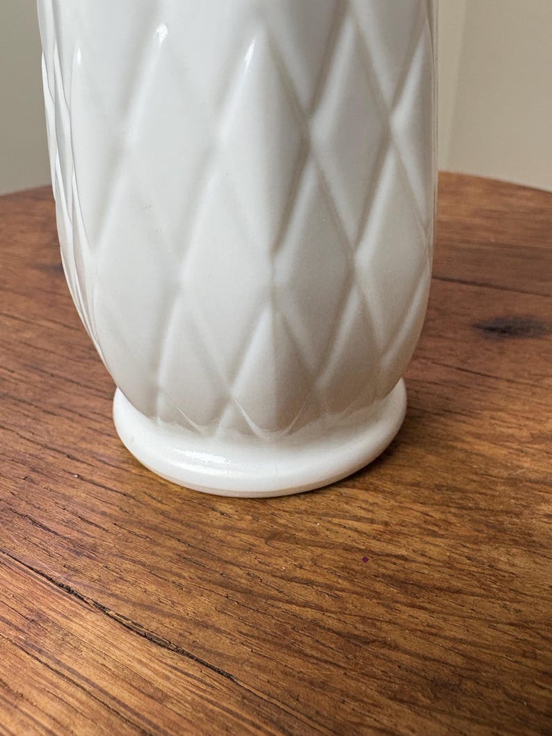 May include: A white ceramic vase with a diamond pattern. The vase has a rounded base and a textured surface. The vase is sitting on a wooden surface.