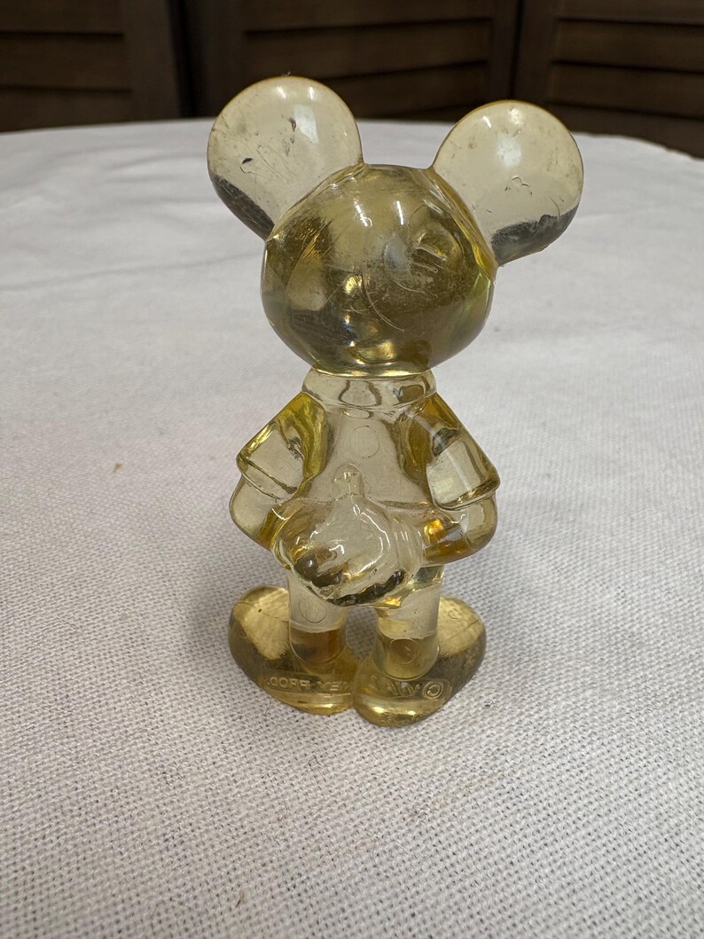 K&ouml;nnte beinhalten: Eine klare, gelblich get&ouml;nte Mickey Mouse-Figur. Die Figur steht, mit gro&szlig;en runden Ohren und einem kleinen Hut. Die Figur besteht aus transparentem Material, der Sockel ist herzf&ouml;rmig.