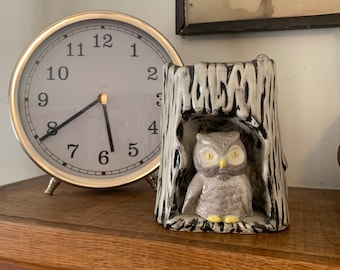 May include: A silver-toned analog clock with a white face and black numbers sits next to a ceramic owl figurine. The owl is gray and yellow, perched inside a tree trunk-shaped holder. The clock is approximately 6 inches in diameter.