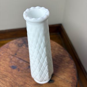 May include: A white, textured vase with a diamond pattern. The vase has a flared, scalloped rim and a cylindrical shape. It is sitting on a round, wooden table.