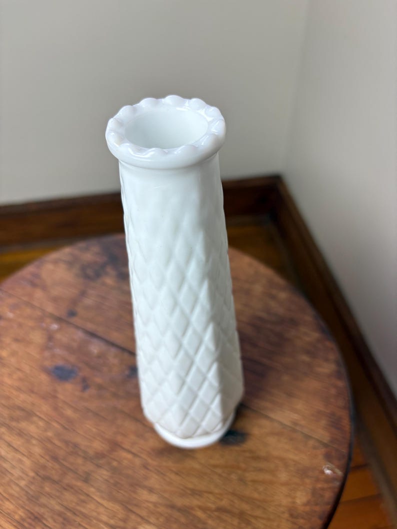 May include: A white, textured vase with a diamond pattern. The vase has a flared, scalloped rim and a cylindrical shape. It is sitting on a round, wooden table.