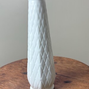May include: A white ceramic vase with a diamond pattern and a scalloped rim. The vase is approximately 9 inches tall and sits on a wooden surface. The vase has a classic, elegant design.