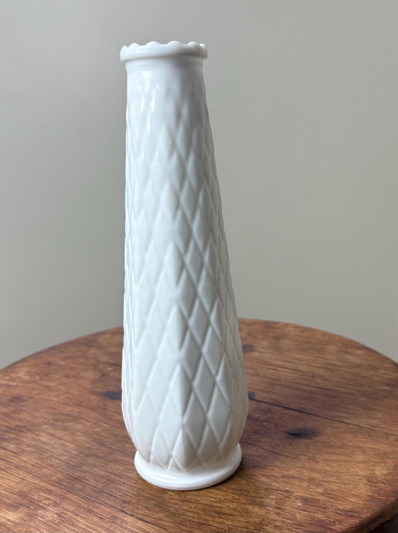 May include: A white ceramic vase with a diamond pattern and a scalloped rim. The vase is approximately 9 inches tall and sits on a wooden surface. The vase has a classic, elegant design.