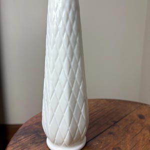 May include: A white, vintage-style vase with a diamond pattern. The vase has a flared top and a round base. The vase is made of a glossy material and is sitting on a wooden surface. The vase is approximately 8 inches tall.