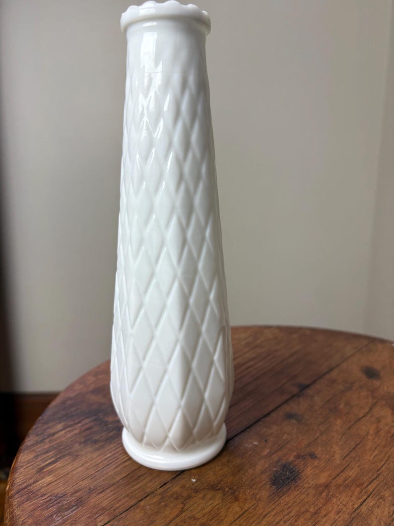 May include: A white, vintage-style vase with a diamond pattern. The vase has a flared top and a round base. The vase is made of a glossy material and is sitting on a wooden surface. The vase is approximately 8 inches tall.
