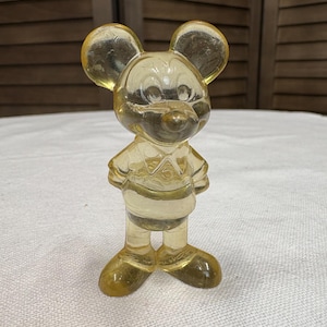 K&ouml;nnte beinhalten: Eine Vintage, durchscheinende gelbe Mickey Mouse Figur. Die Figur steht mit den Armen an den Seiten, tr&auml;gt ein Hemd und Shorts. Die Figur hat gro&szlig;e, runde Ohren und ein l&auml;chelndes Gesicht. Das Material scheint Kunststoff oder Harz zu sein.