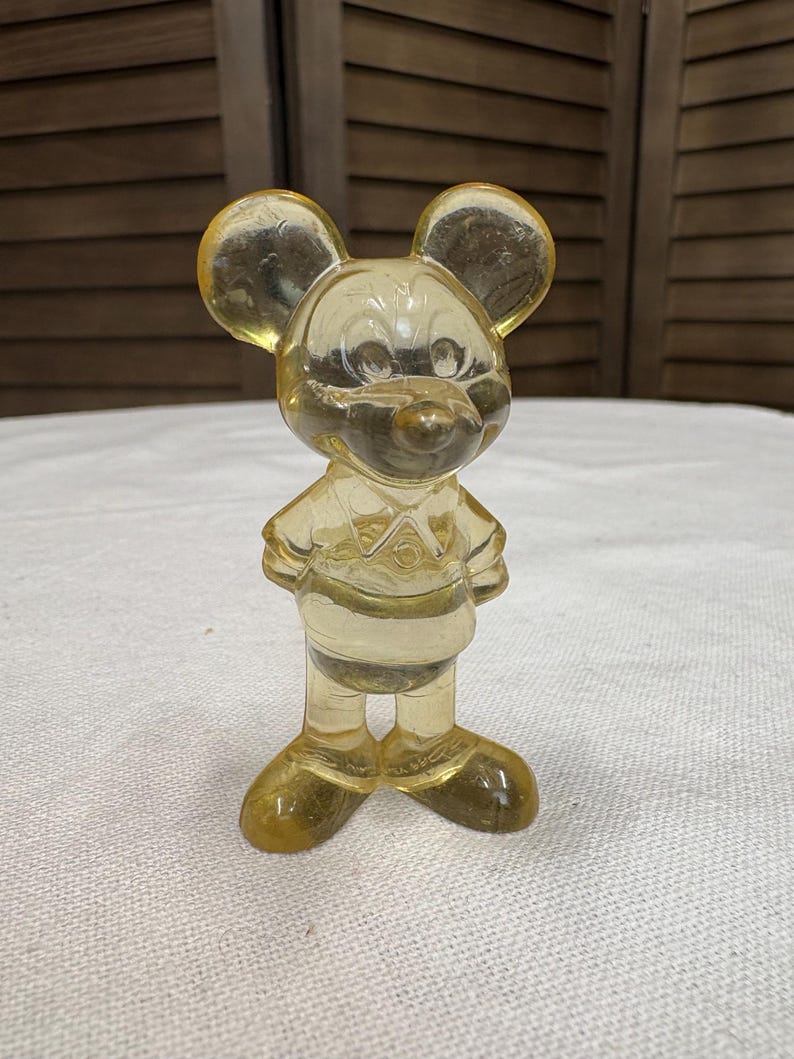 K&ouml;nnte beinhalten: Eine Vintage, durchscheinende gelbe Mickey Mouse Figur. Die Figur steht mit den Armen an den Seiten, tr&auml;gt ein Hemd und Shorts. Die Figur hat gro&szlig;e, runde Ohren und ein l&auml;chelndes Gesicht. Das Material scheint Kunststoff oder Harz zu sein.