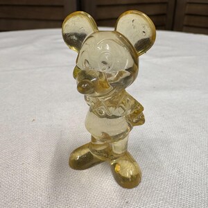 K&ouml;nnte beinhalten: Eine transparente, gelbe Vintage-Figur einer Cartoon-Maus mit gro&szlig;en Ohren, einem runden Gesicht und einem l&auml;chelnden Ausdruck. Die Maus steht aufrecht, die Arme an den Seiten. Die Figur besteht aus einem klaren Material, m&ouml;glicherweise Glas oder Kunststoff.