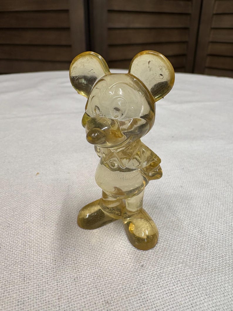 K&ouml;nnte beinhalten: Eine transparente, gelbe Vintage-Figur einer Cartoon-Maus mit gro&szlig;en Ohren, einem runden Gesicht und einem l&auml;chelnden Ausdruck. Die Maus steht aufrecht, die Arme an den Seiten. Die Figur besteht aus einem klaren Material, m&ouml;glicherweise Glas oder Kunststoff.