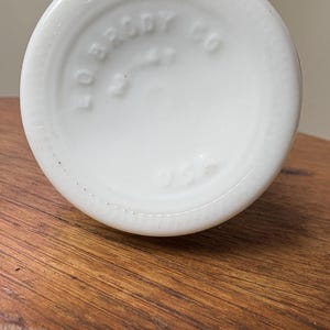 May include: A close-up of the bottom of a white glass bottle. The base is round with the embossed text "EO BRODY CO" and "USA". The bottle sits on a wooden surface.