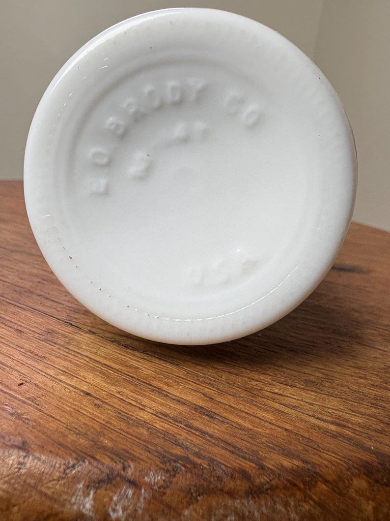 May include: A close-up of the bottom of a white glass bottle. The base is round with the embossed text "EO BRODY CO" and "USA". The bottle sits on a wooden surface.