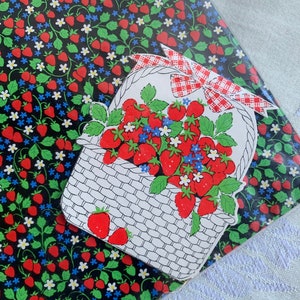 May include: A white basket filled with red strawberries and white flowers with green leaves. The basket has a red and white checkered bow. The basket is on a black background with a repeating pattern of red strawberries and white flowers.