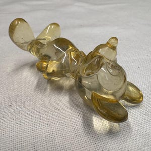 K&ouml;nnte beinhalten: Eine klare, bernsteinfarbene Glasfigur einer Zeichentrickfigur, die auf dem R&uuml;cken liegt. Die Figur hat abgerundete Merkmale, darunter gro&szlig;e Ohren, einen runden K&ouml;rper und ausgestreckte Arme und Beine. Das Glas ist glatt und reflektierend, mit subtilen Farbvariationen.