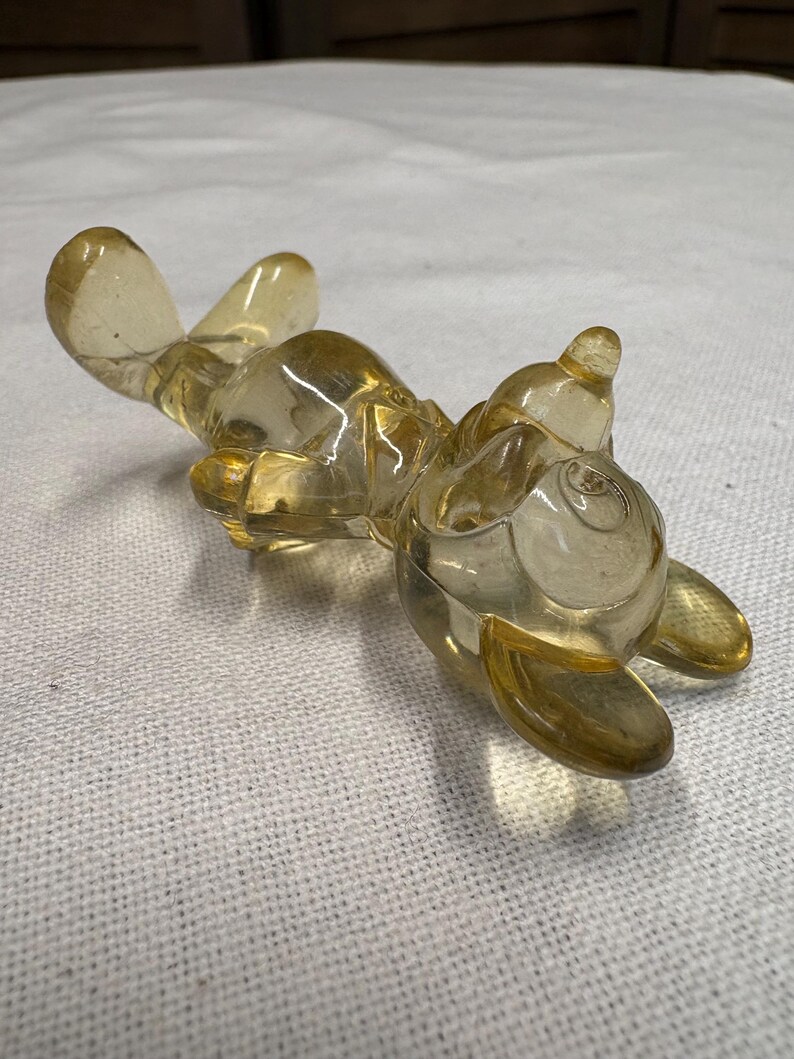 K&ouml;nnte beinhalten: Eine klare, bernsteinfarbene Glasfigur einer Zeichentrickfigur, die auf dem R&uuml;cken liegt. Die Figur hat abgerundete Merkmale, darunter gro&szlig;e Ohren, einen runden K&ouml;rper und ausgestreckte Arme und Beine. Das Glas ist glatt und reflektierend, mit subtilen Farbvariationen.
