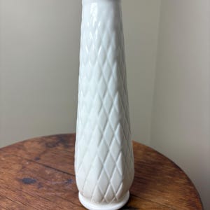 May include: A tall, white vase with a diamond pattern and a scalloped rim. The vase is made of ceramic or porcelain and has a slightly flared base. The vase is approximately 10 inches tall.