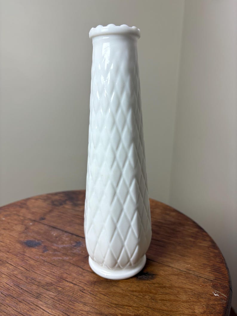 May include: A tall, white vase with a diamond pattern and a scalloped rim. The vase is made of ceramic or porcelain and has a slightly flared base. The vase is approximately 10 inches tall.