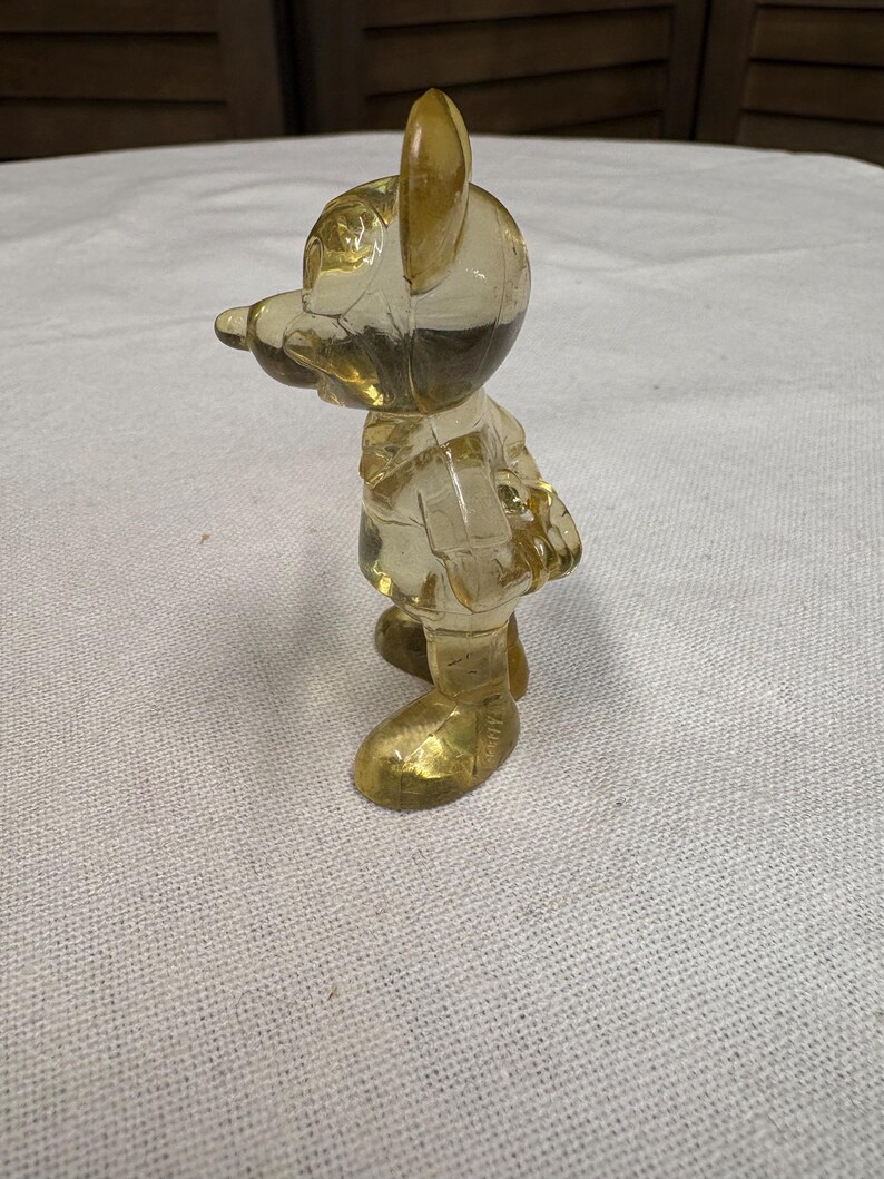 K&ouml;nnte beinhalten: Eine durchscheinende gelbe Figur einer Zeichentrickfigur, die aufrecht steht und die Arme an den Seiten hat. Die Figur hat gro&szlig;e Ohren, eine lange Nase und tr&auml;gt eine Jacke und Schuhe. Die Figur besteht aus einem klaren, m&ouml;glicherweise Kunststoffmaterial.