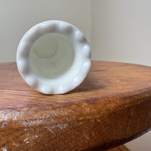 May include: A white, small, vase with a ruffled rim. The vase is made of a glossy material and is set against a neutral background. The vase is resting on a wooden surface.
