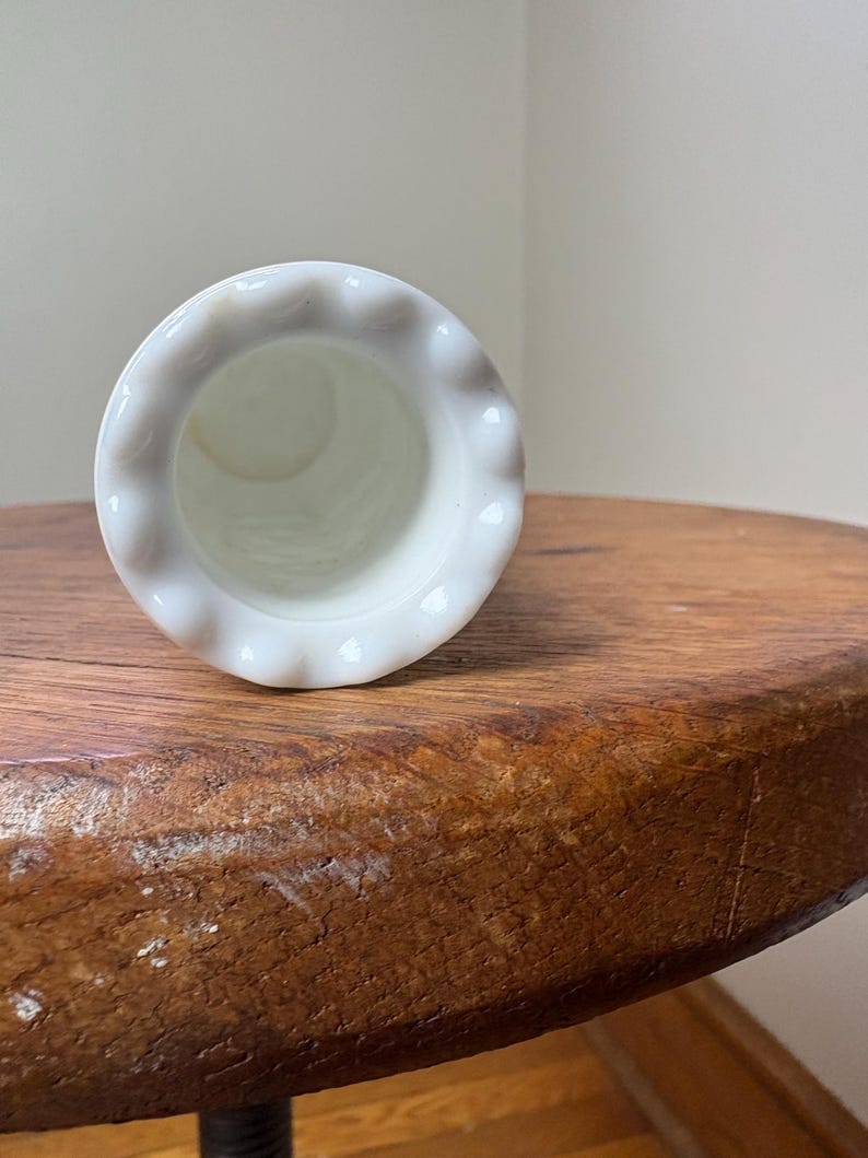 May include: A white, small, vase with a ruffled rim. The vase is made of a glossy material and is set against a neutral background. The vase is resting on a wooden surface.