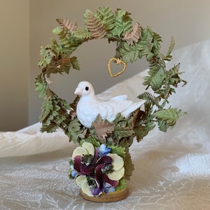 May include: A white porcelain dove perched on a nest of green and brown paper leaves. The nest is on a base decorated with purple, yellow, and white paper flowers. A gold heart hangs from a branch above the dove.