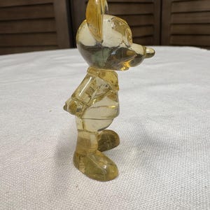 K&ouml;nnte beinhalten: Vintage, transparent gelbe Mickey Mouse Figur. Die Sammlerfigur steht mit den Armen hinter dem R&uuml;cken. Die Figur besteht aus einem durchscheinenden Material, das Licht durchl&auml;sst, und ist vor einem neutralen Hintergrund platziert.