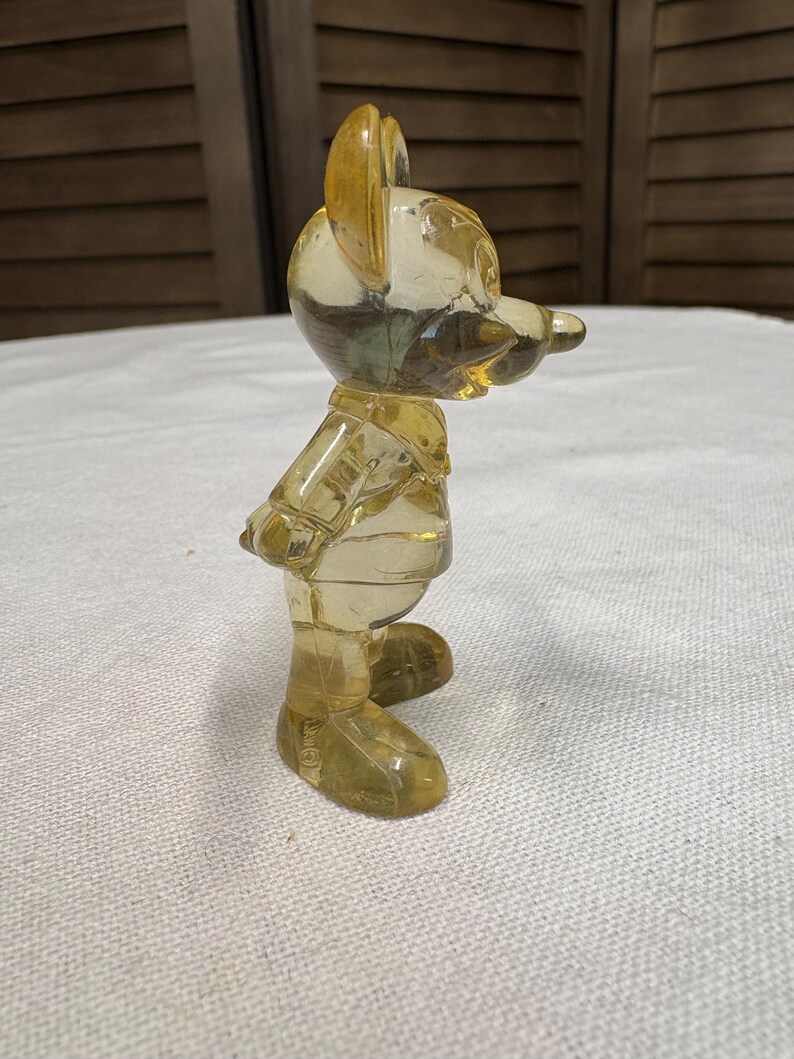 K&ouml;nnte beinhalten: Vintage, transparent gelbe Mickey Mouse Figur. Die Sammlerfigur steht mit den Armen hinter dem R&uuml;cken. Die Figur besteht aus einem durchscheinenden Material, das Licht durchl&auml;sst, und ist vor einem neutralen Hintergrund platziert.