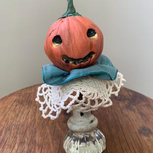 May include: A handmade ceramic pumpkin with a smiling face, wearing a teal blue ribbon and a white crocheted doily. The pumpkin is sitting on a white pedestal with a glass base.