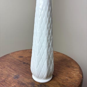 May include: A tall, white vase with a diamond pattern and a ruffled top. The vase is made of ceramic and sits on a wooden surface. The vase is approximately 8 inches tall.
