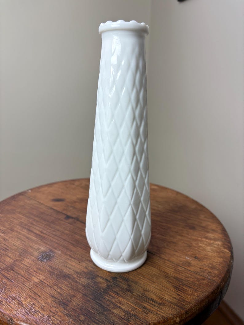 May include: A tall, white vase with a diamond pattern and a ruffled top. The vase is made of ceramic and sits on a wooden surface. The vase is approximately 8 inches tall.