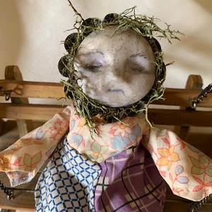 May include: A handmade doll with a patchwork dress and a mossy head. The doll has a sad expression and closed eyes. The doll is sitting on a wooden swing.