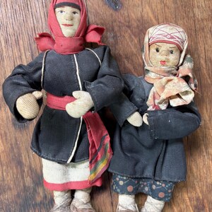 May include: Two handmade dolls dressed in traditional attire. One doll wears a black coat with a red sash and a red hat. The other doll is in a black coat and a patterned headscarf. Both dolls have woven shoes.
