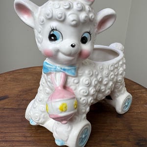 May include: A white ceramic planter shaped like a lamb with a pink bow, blue bow, and a pink rattle. The lamb has blue eyes and a pink nose. The planter has wheels and is on a wooden surface.