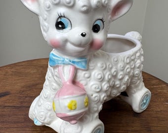 May include: A white ceramic planter shaped like a lamb with a pink bow, blue bow, and a pink rattle. The lamb has blue eyes and a pink nose. The planter has wheels and is on a wooden surface.