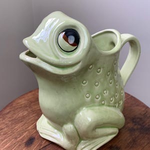 May include: A green ceramic frog pitcher with large, painted eyes. The pitcher has a handle and a spout shaped like a frog's mouth.
