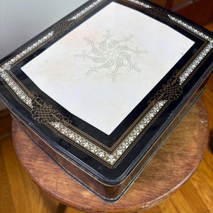 May include: A rectangular black tin box with a white center panel featuring a gold filigree design. The box has a decorative gold and white border. The box is sitting on a small wooden stool.