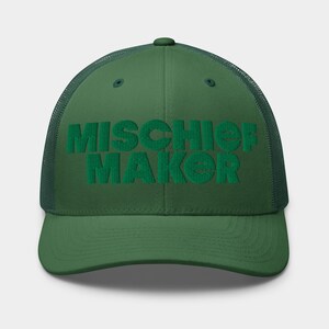 Może przedstawiać: Zielona czapka typu trucker z wyhaftowanym napisem "MISCHIEF MAKER" w pasującym zielonym kolorze. Czapka ma siatkową tylną część i zakrzywiony daszek, oferując klasyczny i swobodny styl. Czapka jest w jednolitym zielonym kolorze.