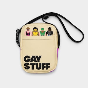 May include: A beige crossbody bag with a black strap and buckle. The bag features the text "GAY STUFF" in large black letters. Above the text are four cartoon figures. The bag has a purple accent on the side.