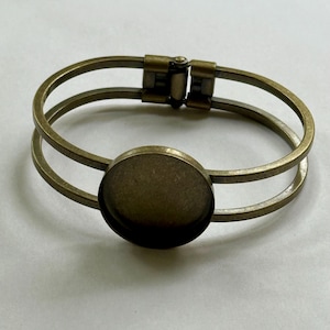 May include: An antique brass-toned bracelet with a double-band design. The bracelet features a round bezel setting in the center, perfect for adding a custom design. The clasp is a hinged style, and the overall aesthetic is vintage.