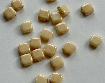 May include: A collection of small, square beads in a creamy beige color. The beads have rounded edges and a slightly glossy finish. They are scattered on a white surface, ideal for jewelry making or crafts.