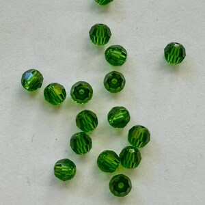 May include: A collection of faceted, emerald green beads. The beads are round with multiple facets that catch the light, creating a sparkling effect. The beads are scattered on a white surface.