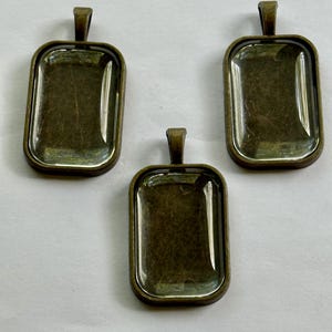May include: Three rectangular pendant trays with a bronze finish. Each tray has a clear glass insert and a small loop at the top for a chain or cord. The trays are empty and ready for customization.