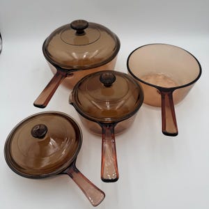 Puede incluir: Juego de cuatro piezas de utensilios de cocina de vidrio color ámbar. Incluye dos cacerolas con tapas y una cacerola sin tapa. La cocina presenta mangos largos integrados y tapas redondas y transparentes con pequeños pomos.