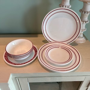 May include: Set of white ceramic dinnerware with red trim. The set includes a large dinner plate, a smaller salad plate, and a bowl. The plates have a simple design with a red band around the edge.